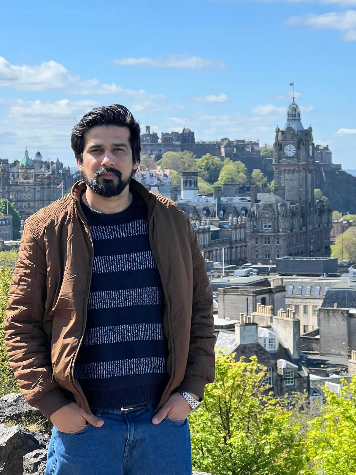 Ahmed Ali Qadir standing on Calton Hill in Edinburgh, with Edinburgh Castle and the Balmoral clock tower behind him