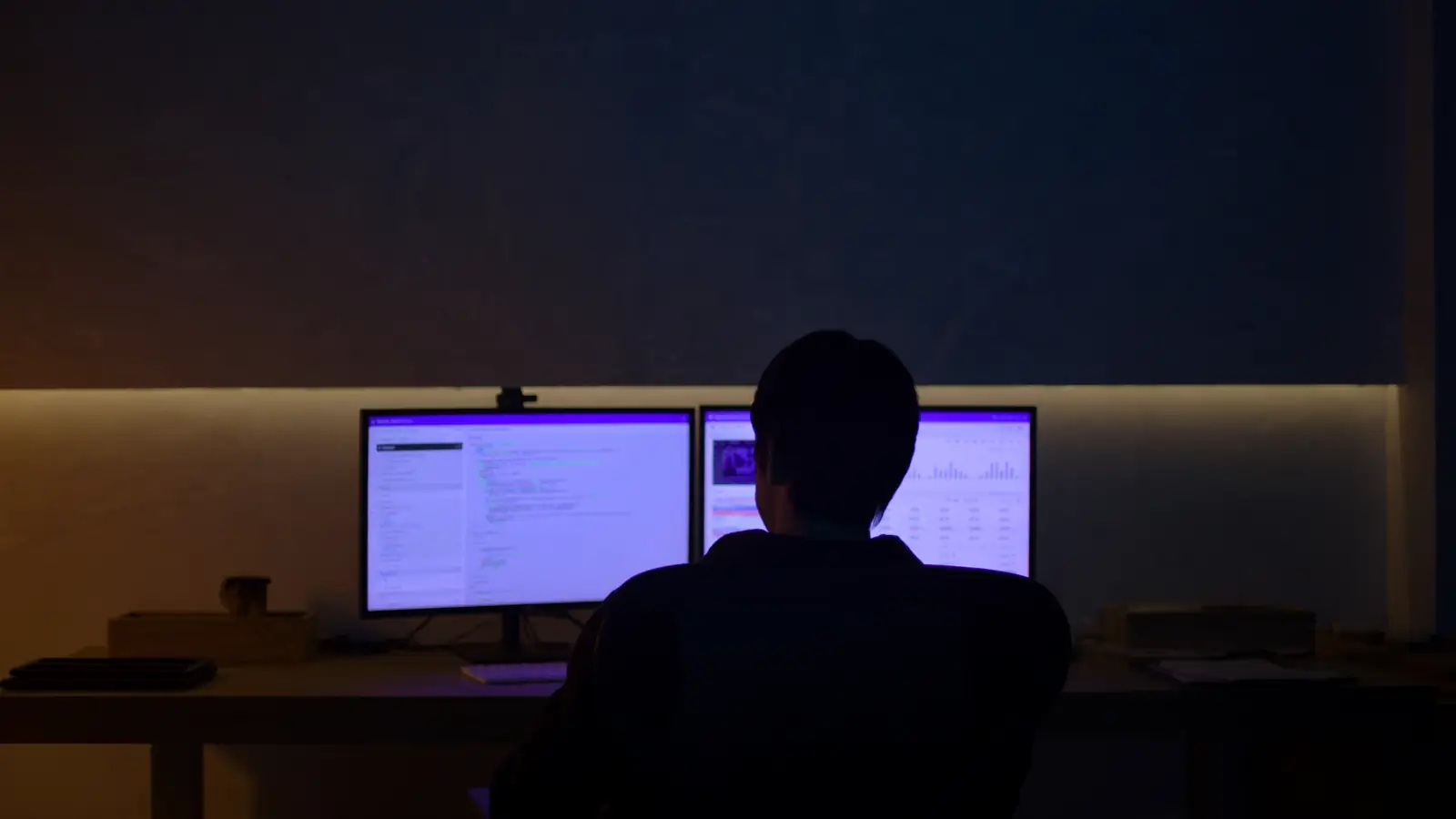 A dark home office at night, a figure seen from behind at a desk lit by two monitors